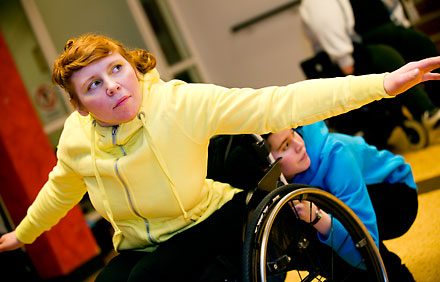 En dansare i gul tröja sträcker ut armarna. En annan dansare hukar bakom hennen rullstol.